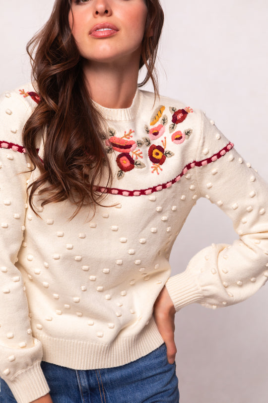 A woman wears the Ivy City Co Sylvie Sweater in Cream, featuring textured polka dots and colorful floral embroidery on the chest, paired with blue jeans. This maternity-friendly style is shown in a photo cropped from shoulders to hips.