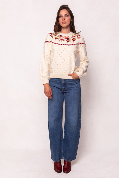A woman poses against a light background in the Ivy City Co Sylvie Sweater in Cream, featuring red floral embroidery. She pairs it with blue wide-leg jeans and red ankle boots for an effortlessly stylish, maternity-friendly look.