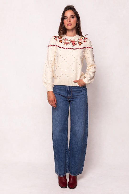 A woman poses against a light background in the Ivy City Co Sylvie Sweater in Cream, featuring red floral embroidery. She pairs it with blue wide-leg jeans and red ankle boots for an effortlessly stylish, maternity-friendly look.