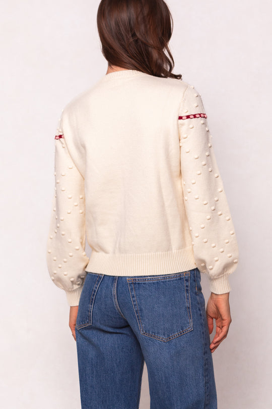 A woman with long brown hair is seen from the back wearing the Ivy City Co Sylvie Sweater in Cream, a maternity-friendly sweater with textured sleeve dots and a red stripe near the shoulders, paired with blue jeans.