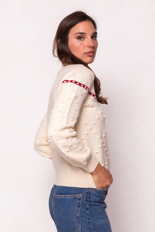 A woman stands sideways, hands in jean pockets, wearing the Ivy City Co Sylvie Sweater in Cream with raised dots and a red stripe. She looks at the camera against a plain light background. This cozy style is maternity friendly.