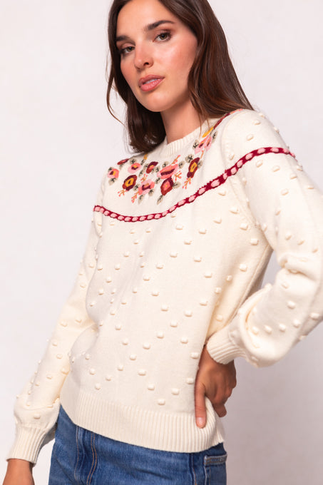 A woman models the Ivy City Co Sylvie Sweater in Cream, featuring textured polka dots, a floral yoke, and a horizontal red ribbon. This maternity-friendly style is paired with blue jeans against a plain light background.