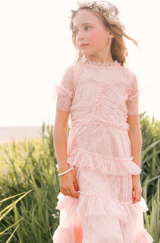 A young girl stands outdoors in the Ivy City Co Mini Whimsical Maxi Dress in Blush, featuring ruffled layers and flutter sleeves. She wears a flower crown and a bracelet, gazing off to the side amid sunlit tall grass.
