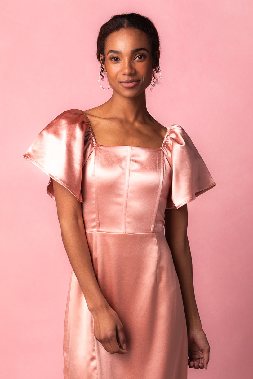 A woman wearing the Ivy City Co Maddie Dress in Apricot Crush Satin, featuring puffed short sleeves, stands against a matching pink background and smiles gently at the camera.
