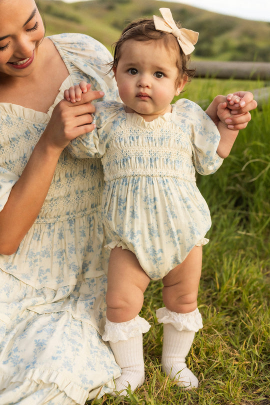 A baby in an Ivory Porcelain Fleur Baby Roselyn Romper by Ivy City Co, featuring a smocked bodice and matching bow, stands on the grass while holding an adult’s hands. Both are dressed in coordinated outfits, with green hills visible in the background.