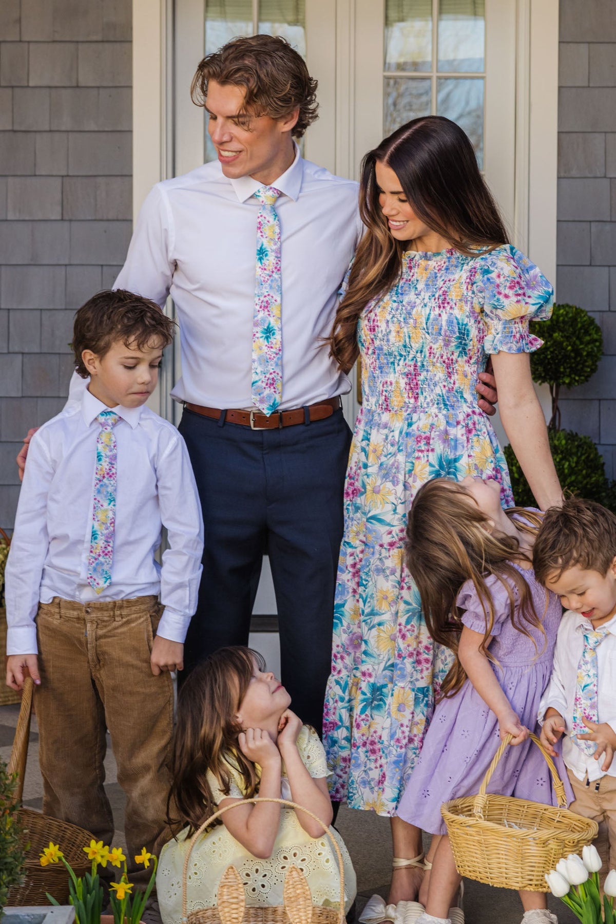 A family of five stands together outside. The adults and two boys wear floral or pastel outfits—with one boy sporting the Ivy City Co Ties in Pretty Plumeria, a cheerful accessory perfect for spring celebrations—while two young girls in dresses hold baskets, possibly for an Easter egg hunt. Yellow flowers bloom in the foreground.