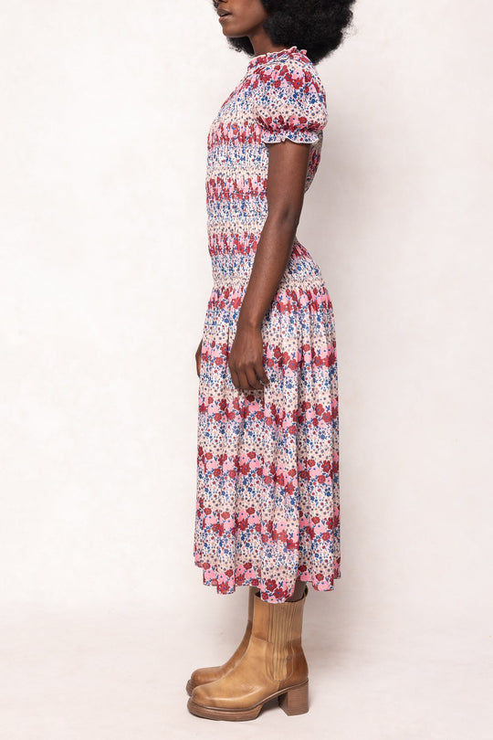 A woman stands in profile wearing the Ivy City Co Hayden Dress in Lush Garden—a short-sleeved, ankle-length floral midi dress featuring red, blue, and white patterns—paired with tan ankle boots. Set against a plain, light-colored background, this maternity-friendly style delivers both comfort and effortless elegance.