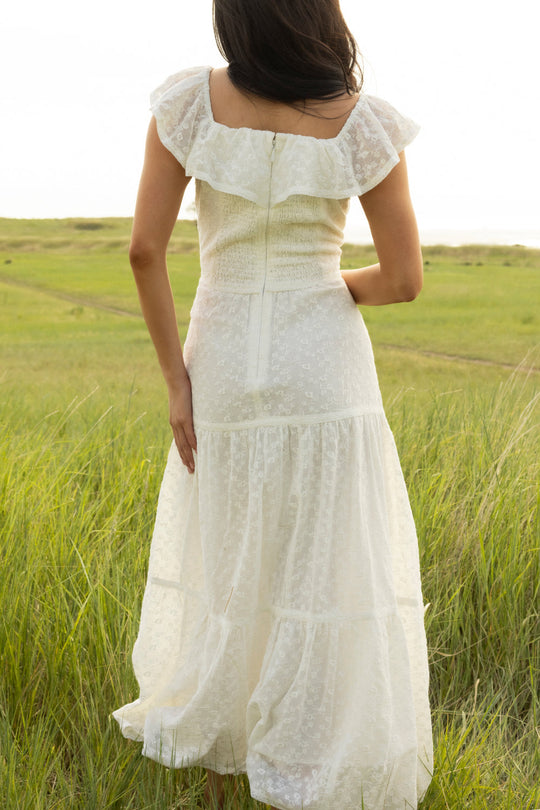 Wearing the Ivy City Co Alaina Dress in Ivory—a white, ruffled, sleeveless maxi dress—a woman stands in a grassy field, facing away from the camera. The scene is bathed in bright, serene natural light.