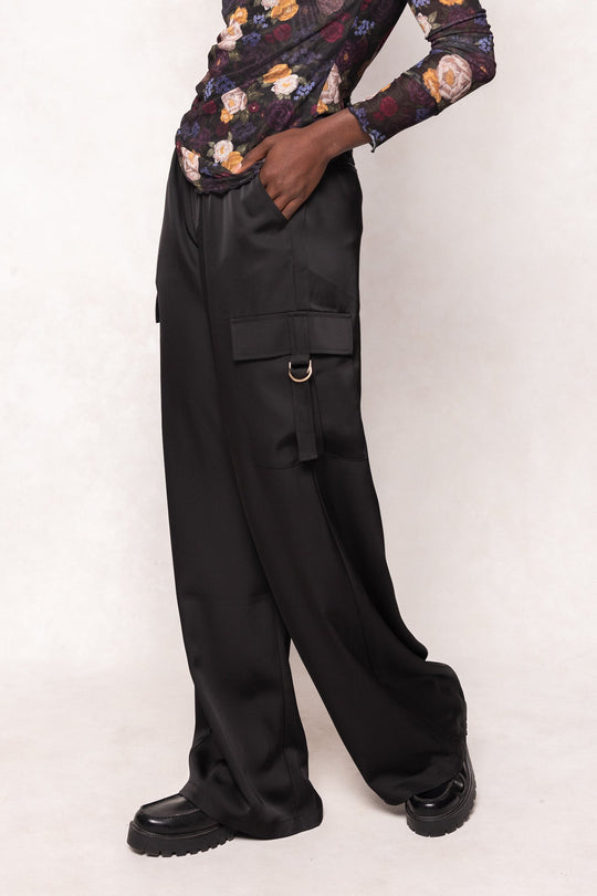 A person stands against a neutral background wearing the Shay Satin Pants in Black by Ivy City Co—loose fit cargo pants with an elastic waistband and stylish ring detail—paired with black platform shoes and a sheer floral top.