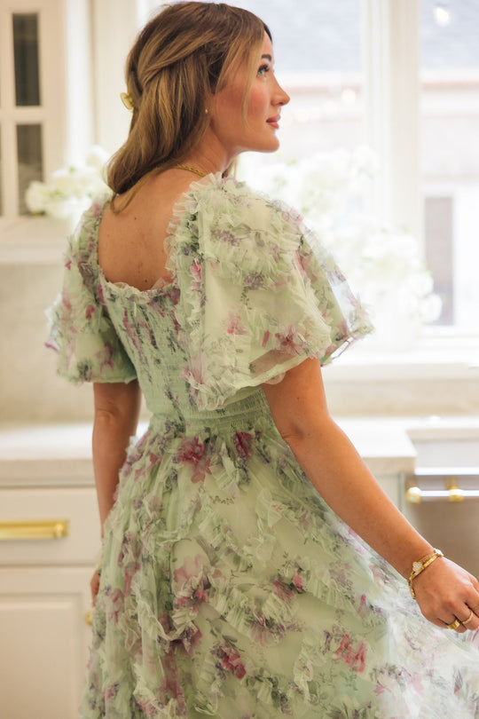 A woman stands sideways in a bright kitchen, sunlight streaming through the window behind her. She wears the Ivy City Co Florentina Dress in Pistachio Aquarelle Fleur—a pastel green, floral, ruffled chiffon piece styled with her light brown hair half up. Designed with maternity-friendly comfort and effortless style, this dress is perfect for new and expectant mothers.