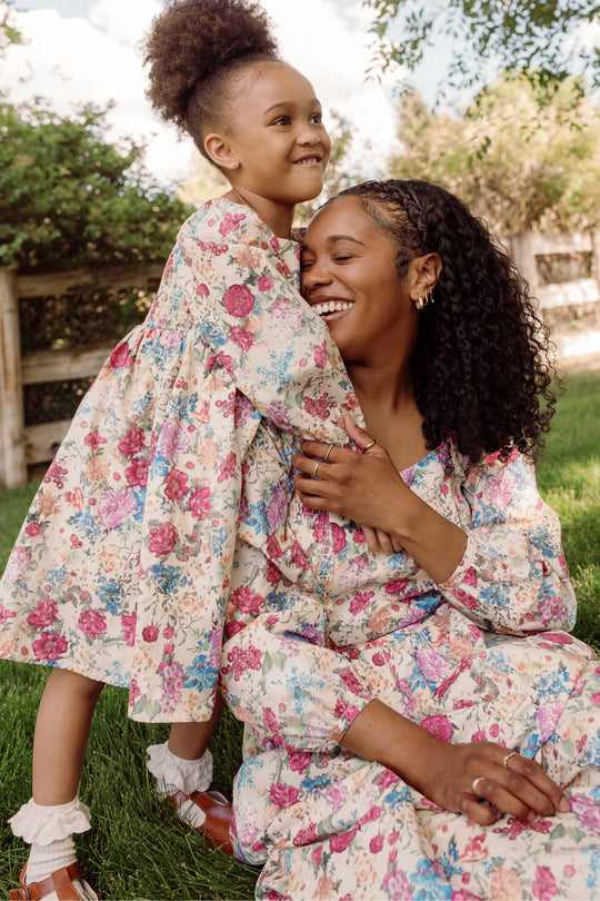 A smiling woman and a young girl, both dressed in matching Ivy City Co Mini Adelpha Dresses in Autumn Amity Floral with elegant balloon sleeves and a delightful smocked bodice, embrace joyfully outdoors on green grass with trees and a wooden fence in the background.