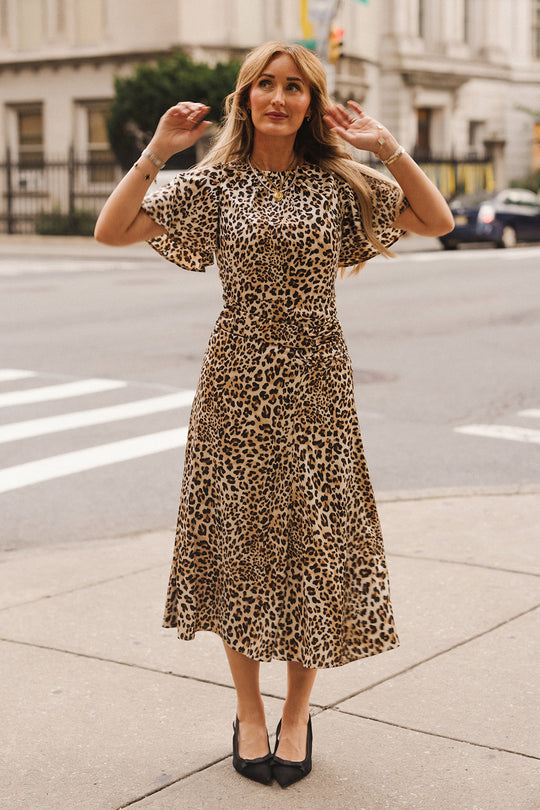 A woman wearing the Tallulah Dress in Leopard by Ivy City Co stands on a city sidewalk, raising her hands near her head. She has long blonde hair and is wearing black heels. Buildings, a crosswalk, and a parked car are visible in the background.