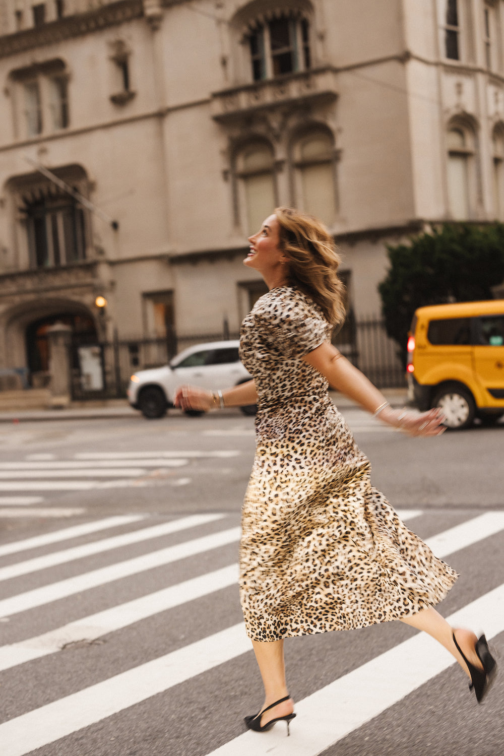 Wearing the Ivy City Co Tallulah Dress in Leopard—with its ruched bodice and flutter sleeves—a woman joyfully walks across a city crosswalk, with cars and a historic building in the background.