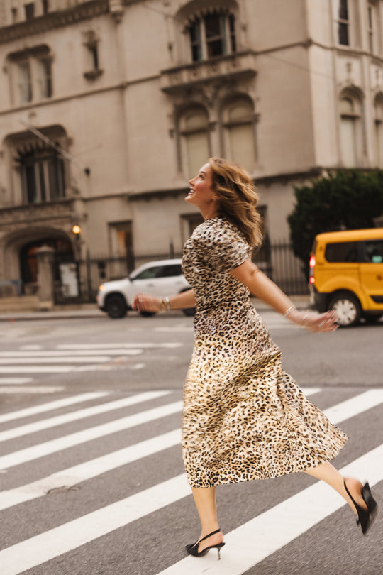 Wearing the Ivy City Co Tallulah Dress in Leopard—with its ruched bodice and flutter sleeves—a woman joyfully walks across a city crosswalk, with cars and a historic building in the background.