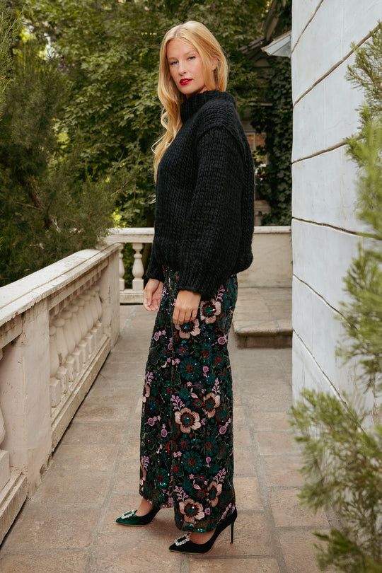 Woman in a black sweater and floral dress standing on a stone path with greenery.