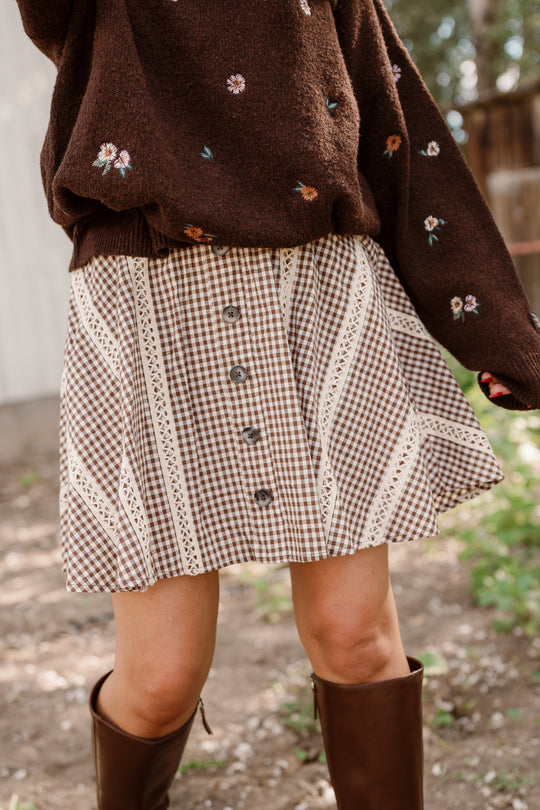 A person stands outdoors on a sunny day wearing a brown floral sweater, the Ivy City Co Tash Skort in Hickory Gingham featuring buttons and lace trim accents, and knee-high brown boots.