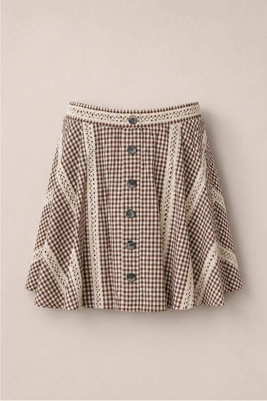 The Tash Skort in Hickory Gingham by Ivy City Co, featuring a brown and white gingham pattern, cream lace trim, front buttons, and an A-line silhouette, is displayed against a light beige background.