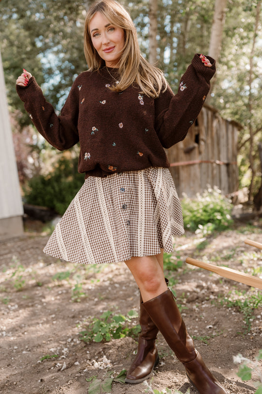 A woman stands outdoors in front of trees and a wooden shed, smiling as she poses with one leg slightly forward. She wears a brown sweater with small floral patterns, knee-high brown boots, and the Ivy City Co Tash Skort in Hickory Gingham—a cotton-blend skort featuring delicate lace trim.