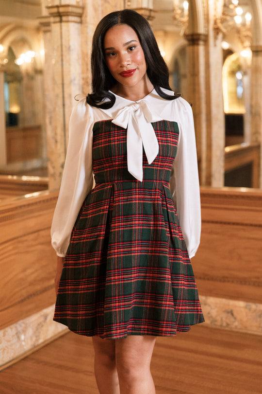A woman stands indoors in an elegant, ornate setting with arched mirrors and warm lighting, wearing the Ivy City Co Tawny Dress in Evergreen Plaid—a perfect girls holiday dress for festive occasions, featuring a classic white blouse with a large bow and a charming evergreen plaid skirt.