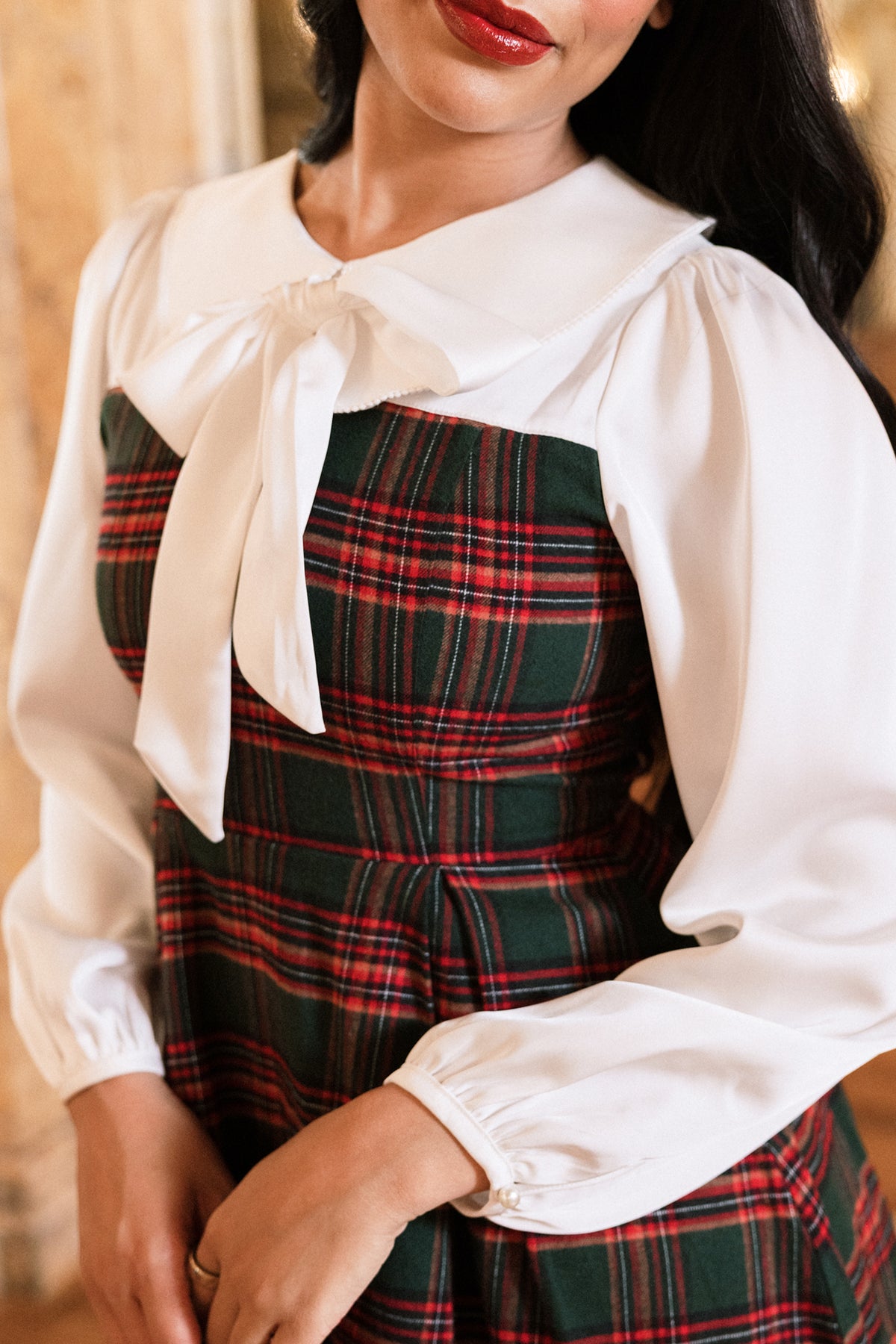 A woman wears the Tawny Dress in Evergreen Plaid by Ivy City Co, featuring long white sleeves, a large white collar, and a bow at the neck. Her dark hair is loosely styled and she has red lipstick. With her hands gently clasped, this dress is perfect for girls during the holidays.