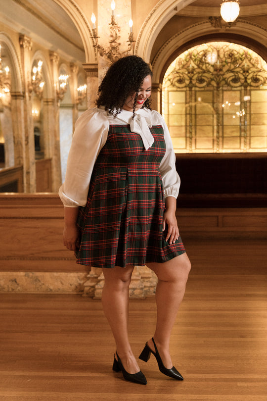Wearing the Ivy City Co Tawny Dress in Evergreen Plaid layered over a white blouse and paired with black heels, a woman stands on a wooden floor in an elegant, warmly lit room adorned with ornate decor and arched mirrors, capturing the timeless charm of this classic Christmas dress.