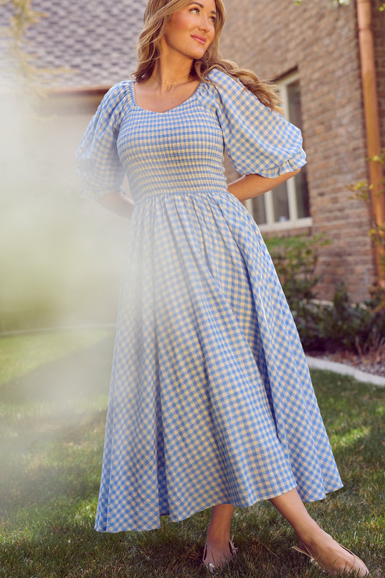 A woman stands on grass near a brick building, wearing the Ivy City Co Teagan Dress in Blue Gingham. The maternity-friendly dress features puffed sleeves. She looks to the side, with one arm relaxed and the other slightly bent.