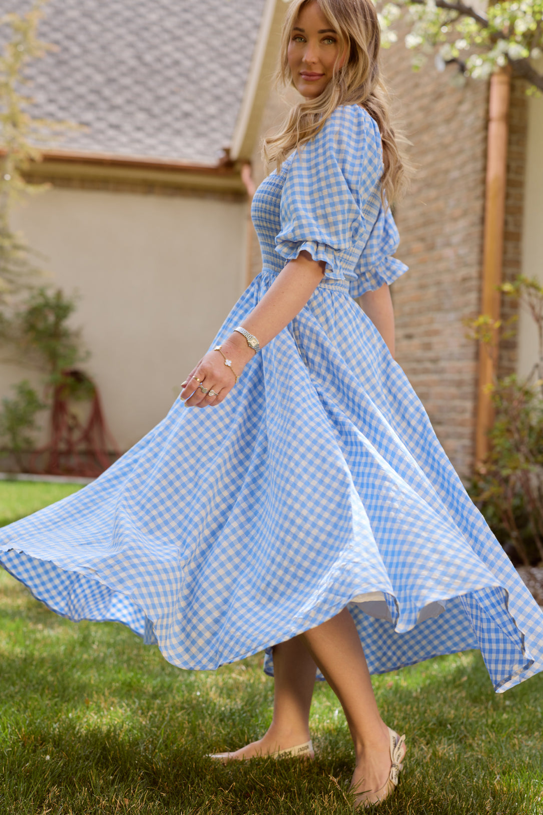 A woman twirls on a grassy lawn in the Ivy City Co Teagan Dress in Blue Gingham, with soft sunlight and a house in the background.