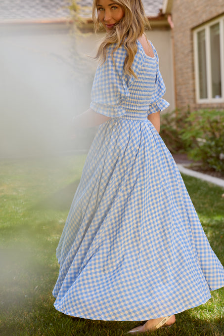 A woman stands outdoors on grass near a building, smiling in the Ivy City Co Teagan Dress in Blue Gingham—a long, nursing-friendly dress that flows out as she turns slightly. A soft blur appears in the foreground.