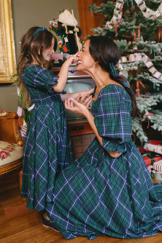 A woman and a young girl, both wearing the Ivy City Co Teagan Dress in Emerald Plaid, smile and play together near a decorated Christmas tree in a warmly lit room. The woman’s dress is designed to be both maternity and nursing friendly, making it perfect for celebrating special moments during the festive season.