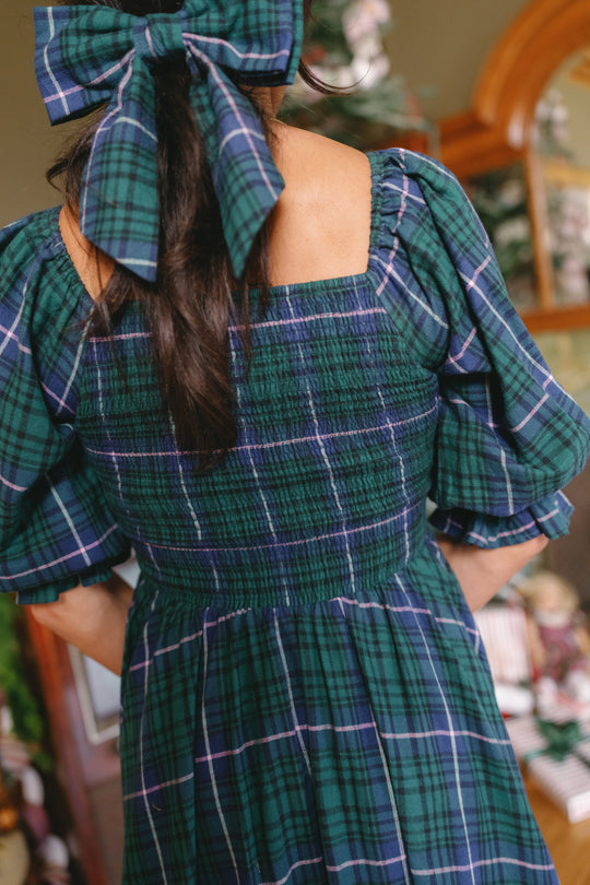 A woman with long dark hair wears the Ivy City Co Teagan Dress in Emerald Plaid, featuring puffed sleeves and a large matching bow, as she stands indoors near a decorated Christmas tree. Its nursing-friendly design makes it perfect for festive gatherings.