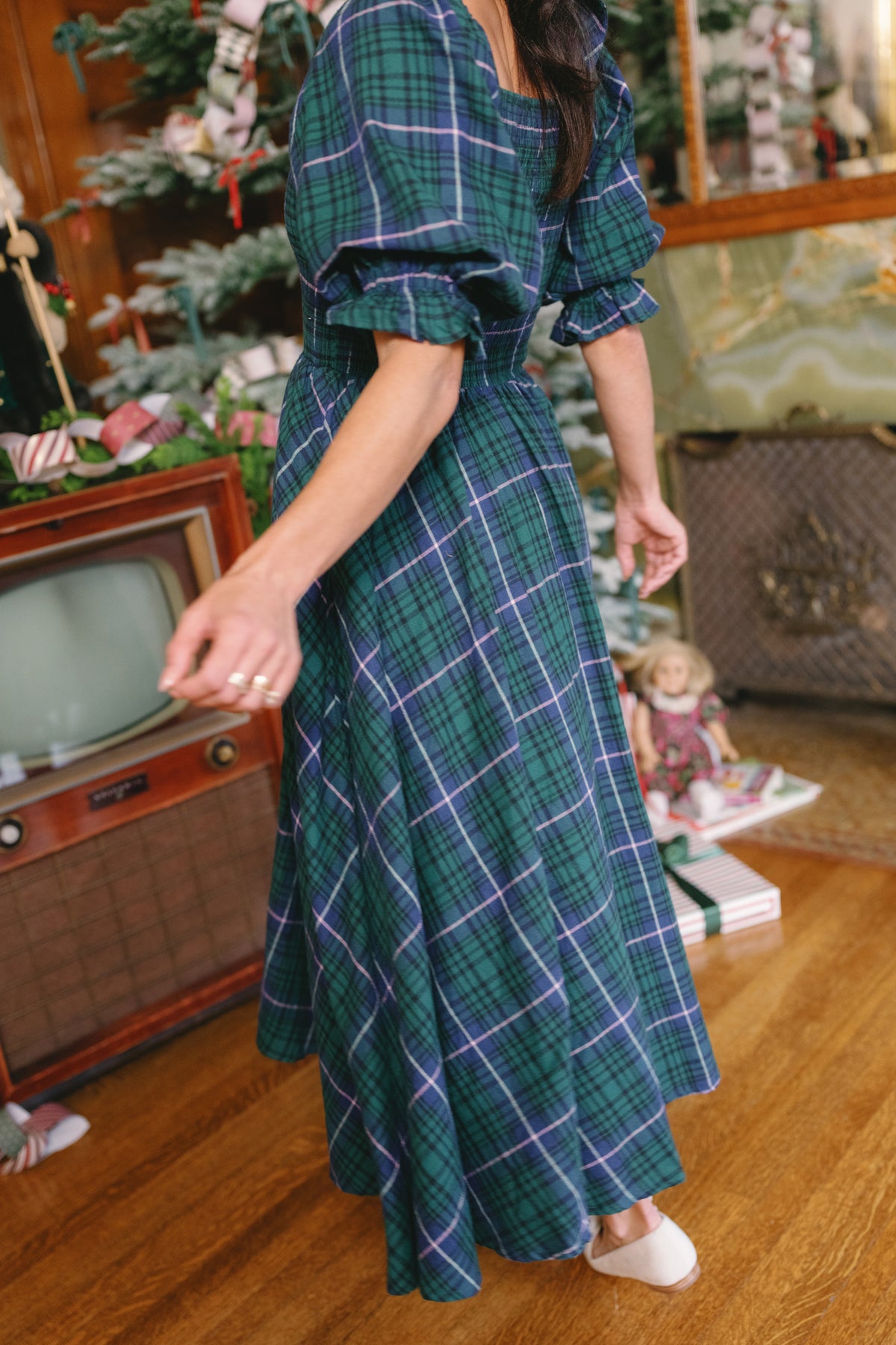 A woman wearing the Ivy City Co Teagan Dress in Emerald Plaid twirls near a vintage TV and a beautifully decorated Christmas tree, surrounded by presents and a doll on the floor. This joyful scene embodies holiday spirit with a festive, nursing-friendly design perfect for new mothers.