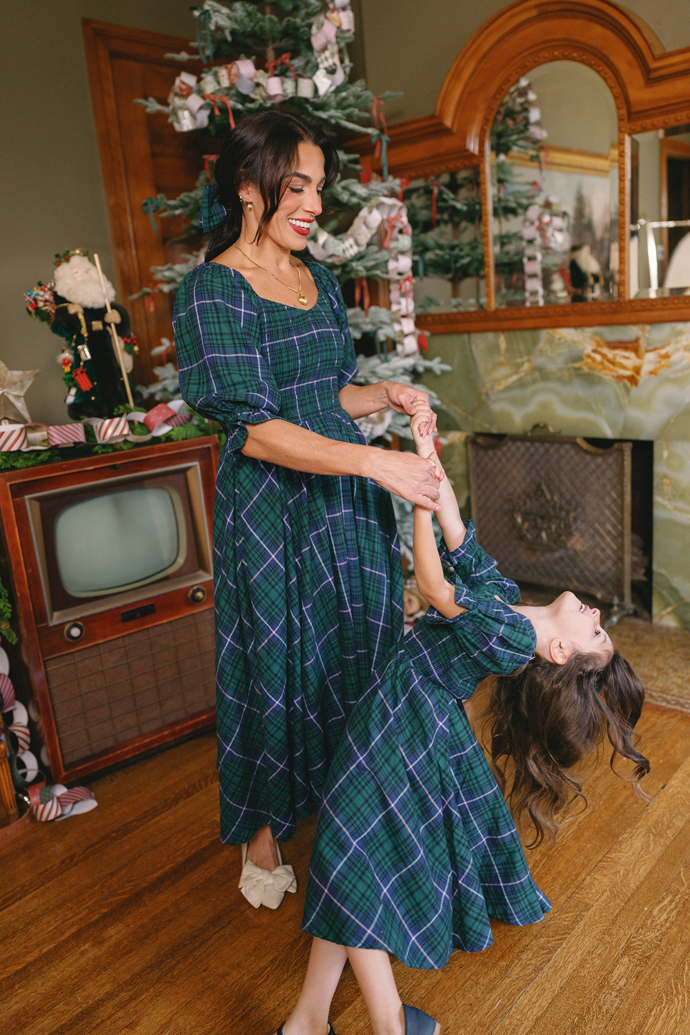 A woman and a young girl, both wearing the Ivy City Co Teagan Dress in Emerald Plaid, smile and dance together near a vintage TV and decorated Christmas tree in a cozy living room.