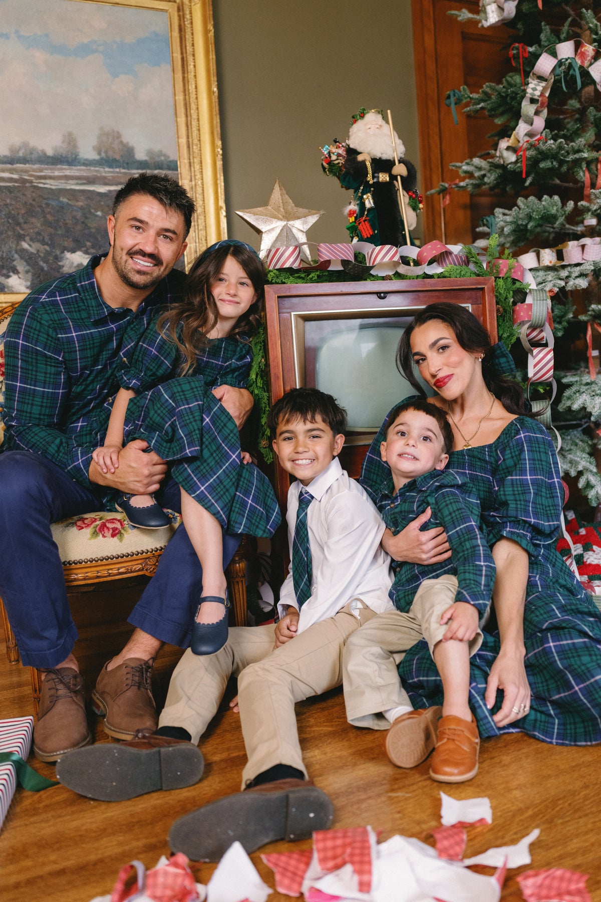 A family of five in matching plaid outfits sits near a Christmas tree and vintage TV, surrounded by presents and festive decorations, smiling warmly at the camera. The mother glows in the Ivy City Co Teagan Dress in Emerald Plaid—a cozy, nursing-friendly holiday dress that makes this special moment both stylish and comfortable for everyone.