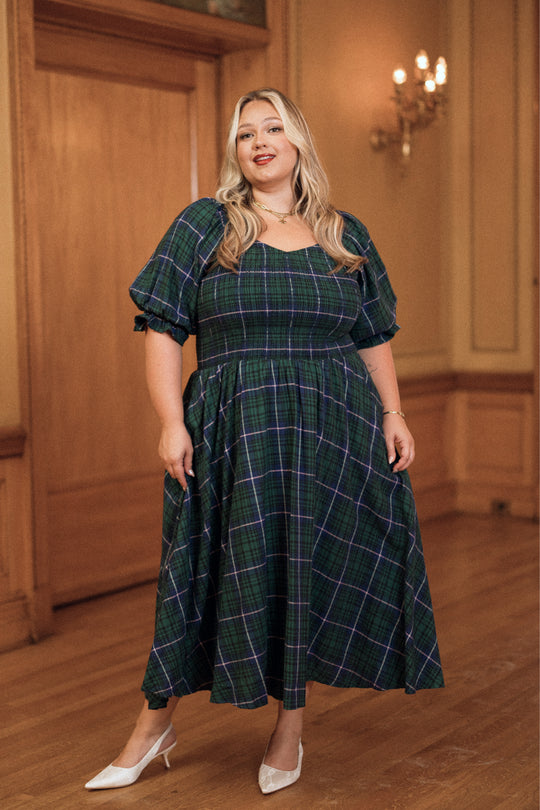 A woman in the Ivy City Co Teagan Dress in Emerald Plaid—a green and blue plaid maternity dress with puff sleeves—stands in an elegant wood-paneled room, smiling and wearing white heels. This stylish, nursing-friendly dress is a perfect holiday choice for mothers-to-be.