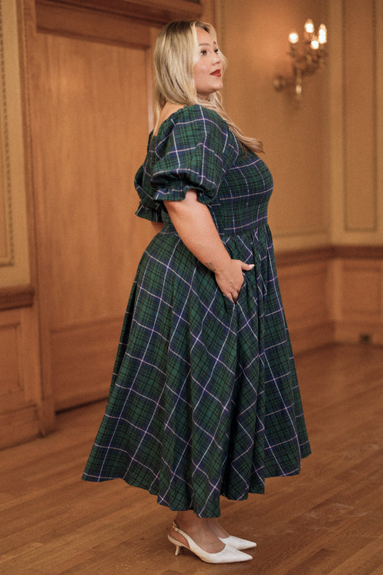 A woman stands indoors wearing the Teagan Dress in Emerald Plaid by Ivy City Co, featuring puffed sleeves, styled with white heels as she looks to the side. The room has wooden floors, paneling, and a chandelier in the background.