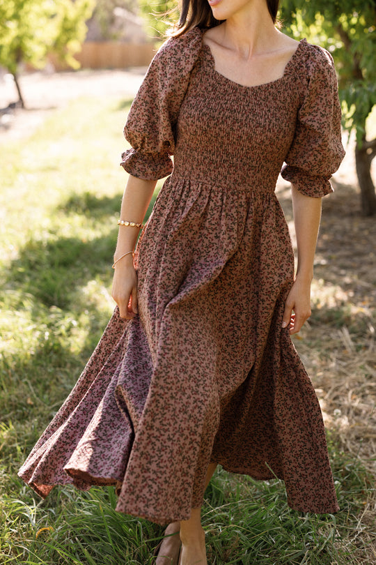 Wearing the Ivy City Co Teagan Dress in Mauve Woodland Floral, a woman stands on grass outdoors, her figure partially turned as sunlight creates soft shadows. The long dress features puffed sleeves and a smocked bodice—an elegant style that’s also maternity friendly.