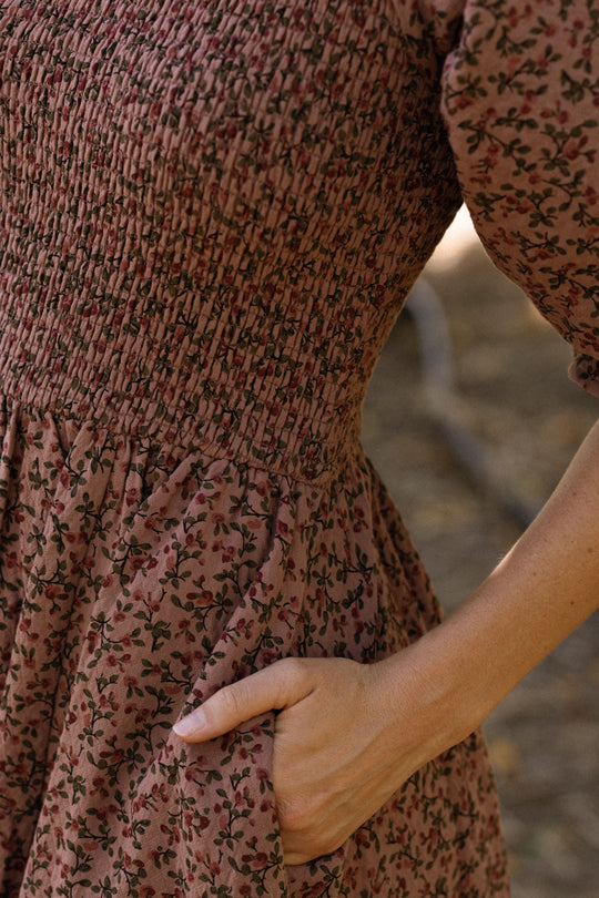 A person wearing the Ivy City Co Teagan Dress in Mauve Woodland Floral—a brown, floral-patterned maternity-friendly style with a smocked bodice and puffy sleeve—stands outdoors, one hand slipped into a side pocket. Only the torso and part of the arm are visible.