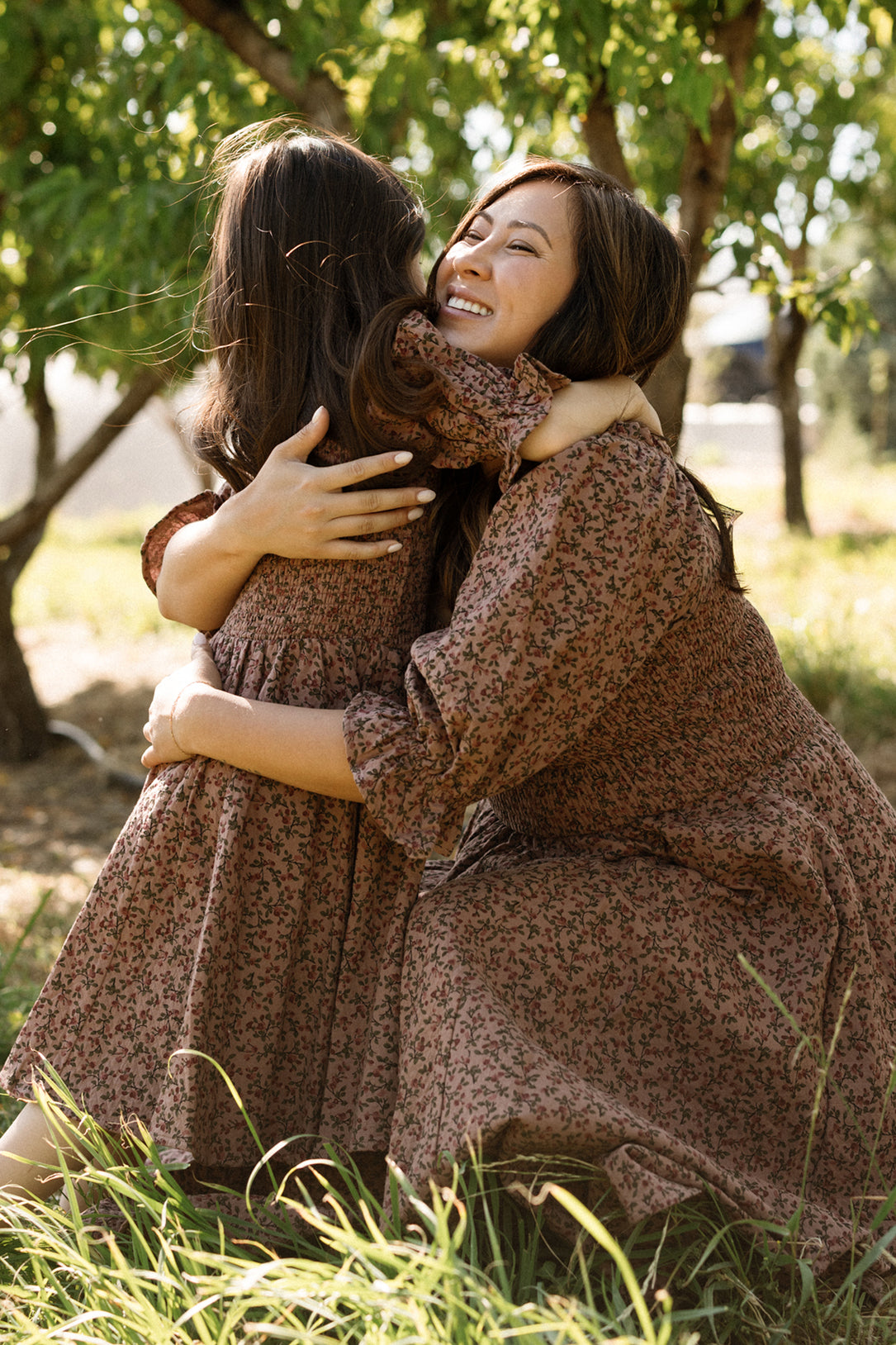 A mother and daughter, both wearing matching Ivy City Co Teagan Dresses in Mauve Woodland Floral with smocked bodices, hug each other joyfully outdoors in a sunny, green park—the maternity-friendly style making their moment even more special.