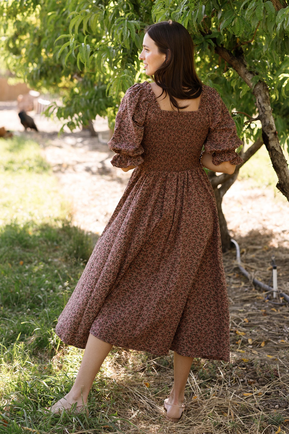 A woman stands outdoors on the grass, facing away, wearing the Ivy City Co Teagan Dress in Mauve Woodland Floral. Sunlight filters through the leafy branches of a nearby tree, highlighting the dress’s smocked bodice and maternity-friendly design.