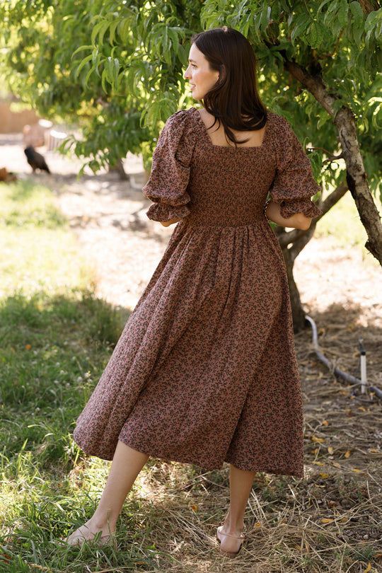 A woman stands outdoors on the grass, facing away, wearing the Ivy City Co Teagan Dress in Mauve Woodland Floral. Sunlight filters through the leafy branches of a nearby tree, highlighting the dress’s smocked bodice and maternity-friendly design.