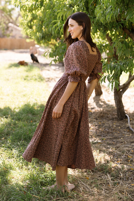 A woman smiles outdoors on a grassy lawn near green trees, sunlight dappling through the leaves, as she stands to the side wearing Ivy City Co’s Teagan Dress in Mauve Woodland Floral. The dress features a smocked bodice and flat shoes, offering both style and maternity-friendly comfort.