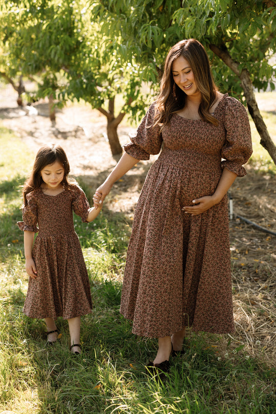 A pregnant mother with long brown hair walks through a sunlit orchard, smiling down as she holds hands with her young daughter. Both are wearing matching Ivy City Co Teagan Dresses in Mauve Woodland Floral, featuring smocked bodices designed to be beautifully maternity friendly. The mother’s hand rests gently on her baby bump as they stroll together.