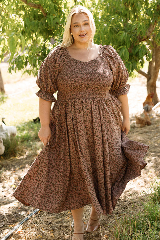 A woman stands outdoors in the Teagan Dress in Mauve Woodland Floral by Ivy City Co, holding her skirt and smiling. Sunlight filters through green trees in the background, creating a warm and natural ambiance. The dress features a smocked bodice, is maternity friendly, and perfectly blends comfort with style for enjoying moments amidst nature.