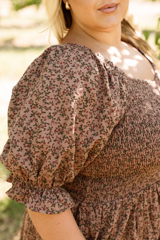 A woman wearing the Ivy City Co Teagan Dress in Mauve Woodland Floral stands outdoors in sunlight, revealing only her torso, arm, and part of her face, against a softly blurred natural background. The dress features puff sleeves and a smocked bodice, making the style beautifully maternity friendly.