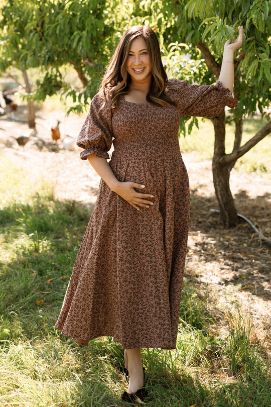 A woman wearing the Ivy City Co Teagan Dress in Mauve Woodland Floral, featuring a smocked bodice and maternity-friendly design, stands outdoors beneath a tree. She smiles gently, one hand resting on her belly and the other holding a branch, as sunlight filters through the leaves onto the grass.