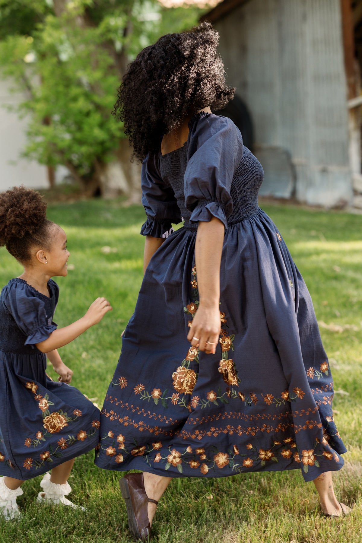 A woman and a young girl run and play on the grass outdoors, both wearing matching Ivy City Co Teagan Dress in Navy Embroidered Fall Floral—with stylish, maternity-friendly smocked bodices—set against a backdrop of trees and a rustic building.