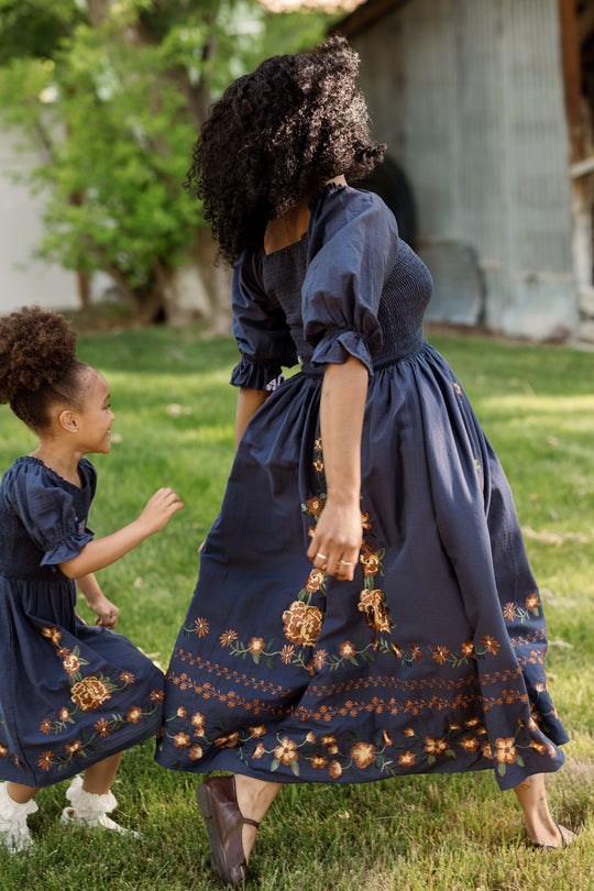 A woman and a young girl run and play on the grass outdoors, both wearing matching Ivy City Co Teagan Dress in Navy Embroidered Fall Floral—with stylish, maternity-friendly smocked bodices—set against a backdrop of trees and a rustic building.