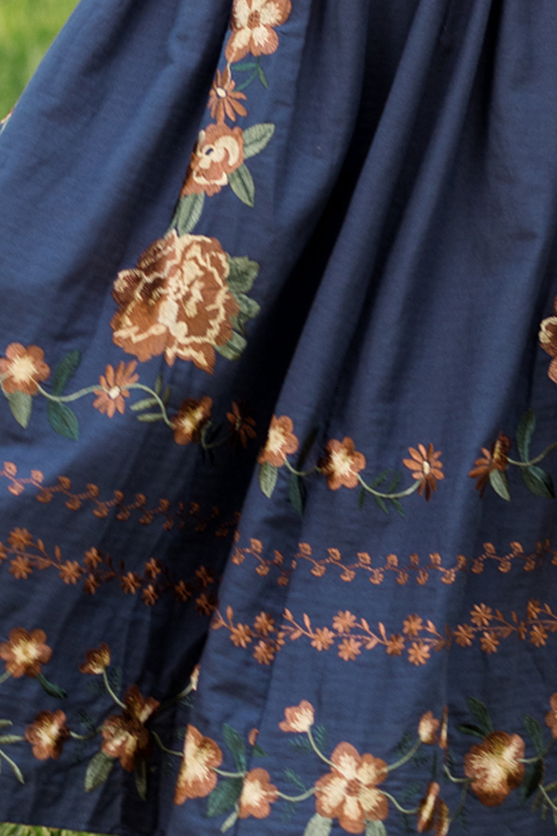 A close-up of the Ivy City Co Teagan Dress in Navy Embroidered Fall Floral, showcasing intricate orange and green floral embroidery along the hem and lower part of the fabric, with a smocked bodice for a comfortable, maternity-friendly fit.