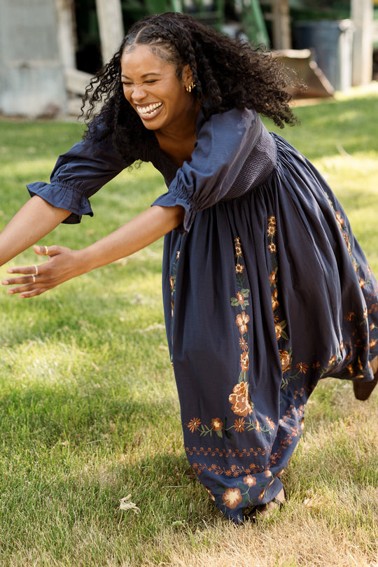 A woman wearing the Ivy City Co Teagan Dress in Navy Embroidered Fall Floral—with its flowing silhouette, smocked bodice, and detailed floral embroidery—laughs joyfully with outstretched arms while standing outdoors on lush green grass. This elegant style is also maternity friendly.