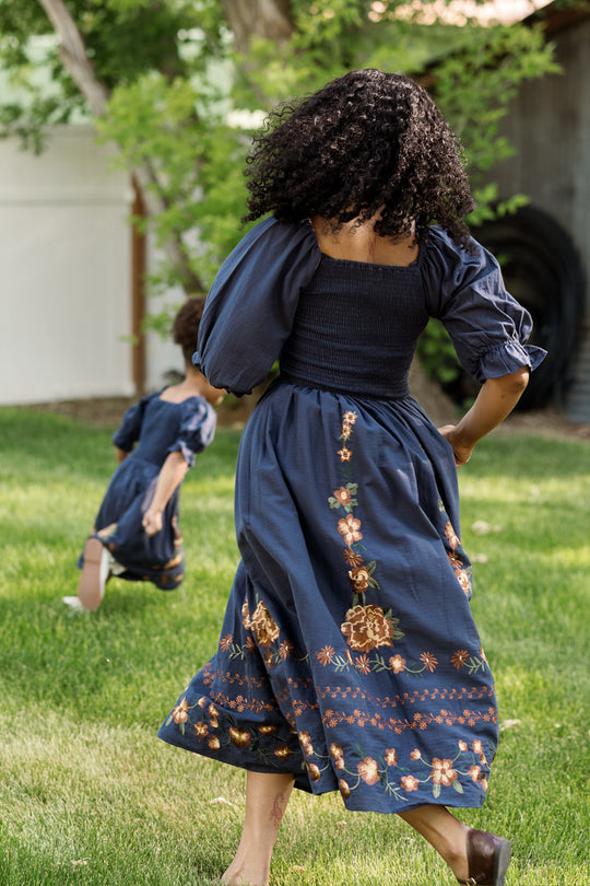 A woman and a child, both wearing Ivy City Co’s Teagan Dress in Navy Embroidered Fall Floral—with delicate floral patterns and smocked bodice details—run barefoot across green grass in a sunny backyard. Their comfy matching Ivy City Co dresses are perfect for play, with trees and a white fence completing the scene in the background.